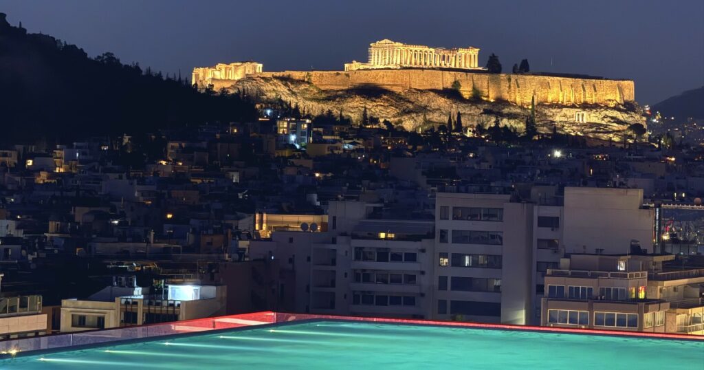 Illuminated Acropolis overlooking Athens cityscape with turquoise rooftop pool in foreground.