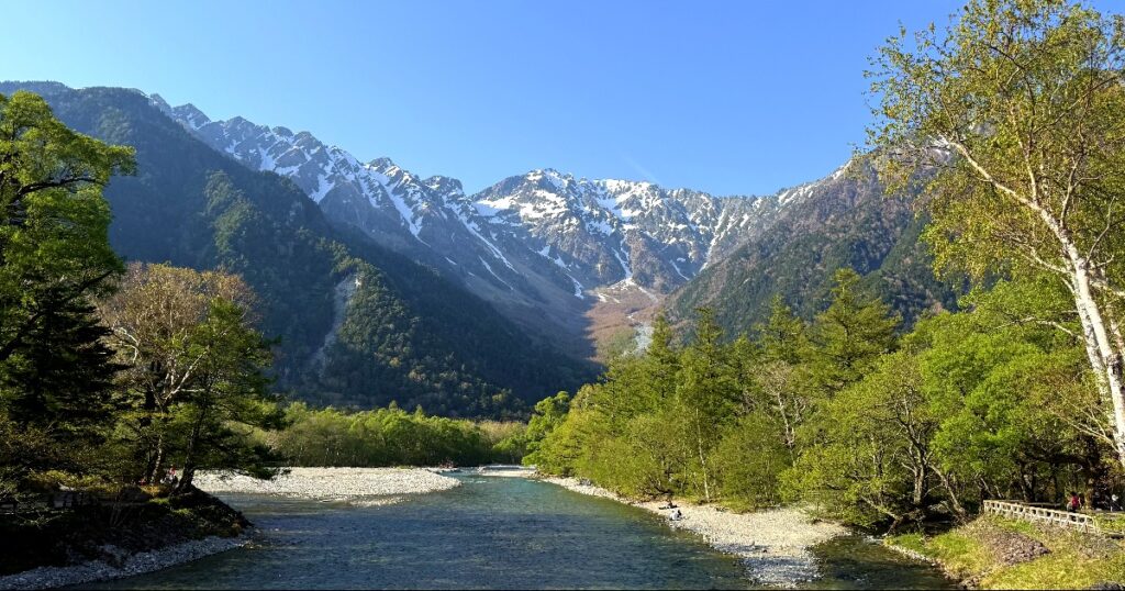 Crystal clear river flows through lush valley surrounded by snow-capped alpine mountains and forest in Kamikochi.
