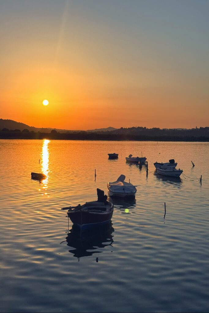 Silhouetted fishing boats anchored in calm golden sunset waters of a Greek island harbor.