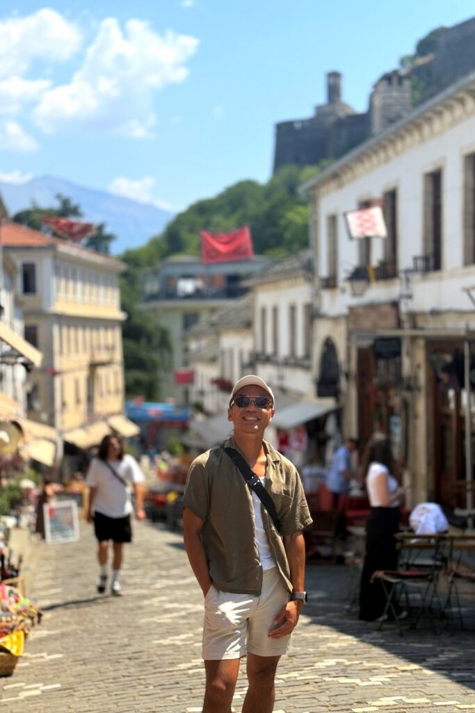 Man walking through historic cobblestone street in Gjirokaster with castle overlooking the mountainous Albanian town.