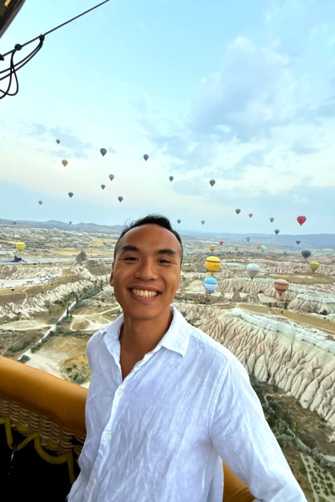 Young man smiling in hot air balloon above Cappadocia's fairy chimneys and landscape.