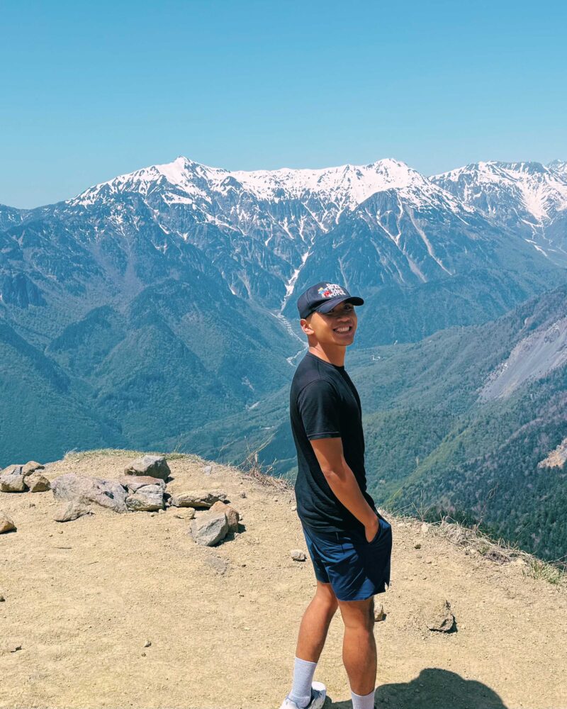 Traveler standing on a mountain summit with snow-capped peaks and valley views in Kamikochi Japan.