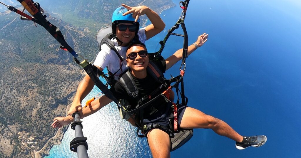 Two paragliders soaring above mountains and turquoise Mediterranean waters in Ölüdeniz, Turkey.