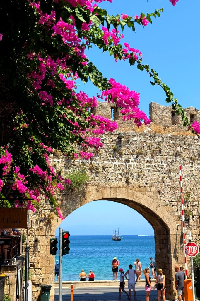 Vibrant pink bougainvillea flowers frame historic stone archway overlooking Mediterranean Sea in Rhodes, Greece.