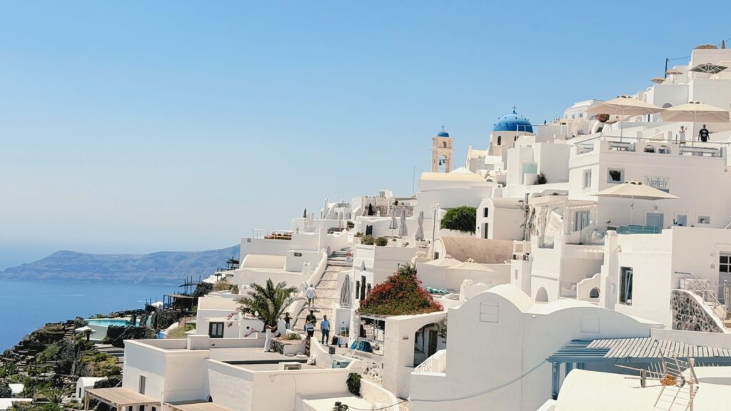 Bright midday view of Oia, Santorini, with white-washed Cycladic buildings cascading toward the caldera, featuring a blue-domed church, bell tower, and the deep blue Aegean Sea in the background