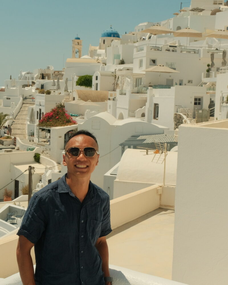 Traveler posing in Oia, Santorini, with the iconic white-washed buildings and blue Aegean Sea in the background.