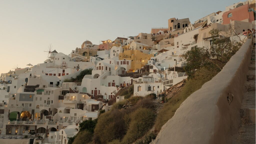 White-washed buildings and windmills cascading down the hillside of Oia, Santorini, photographed at dusk with warm golden light illuminating the terraced Cycladic architecture