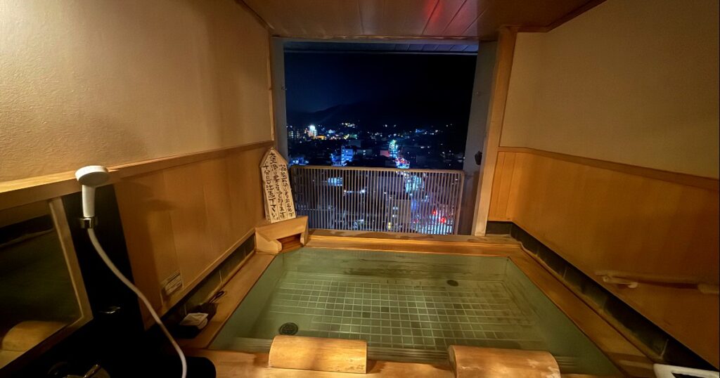 Traditional Japanese onsen hot spring bath with nighttime city lights visible through large window.