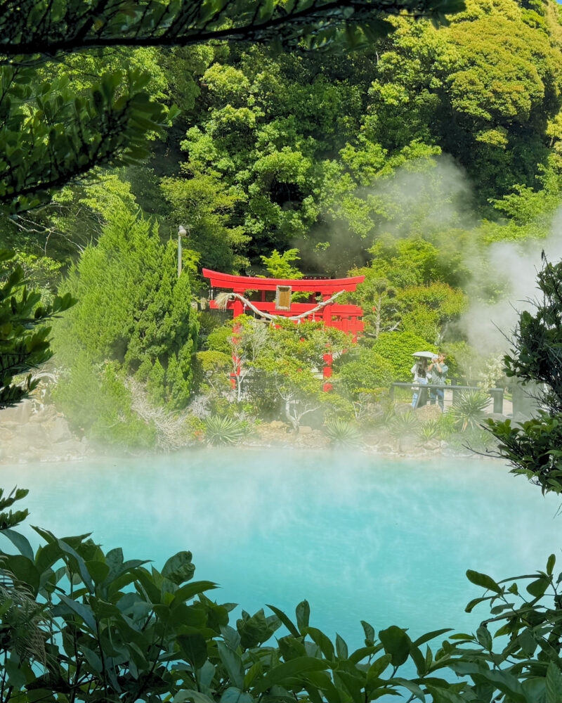 Red bridge spanning turquoise hot spring surrounded by lush green forest at Umi Jigoku, Beppu.