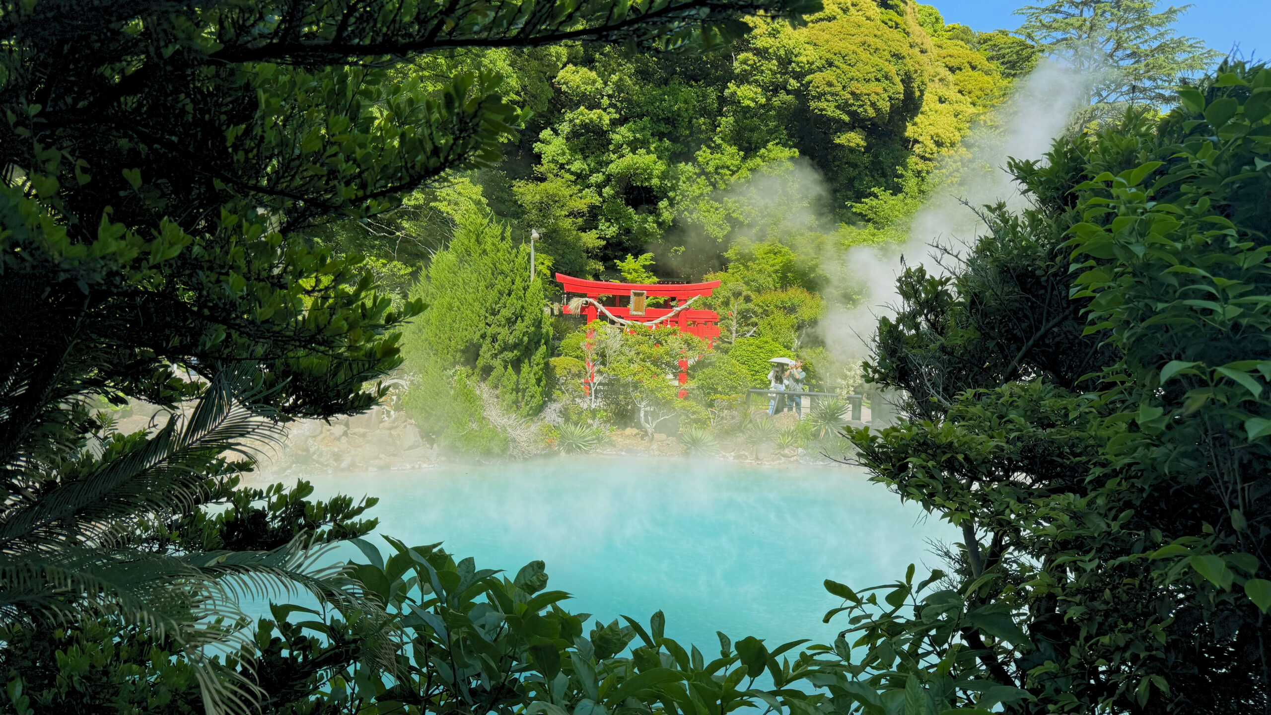 Red bridge spanning turquoise hot spring surrounded by lush green forest at Umi Jigoku, Beppu.