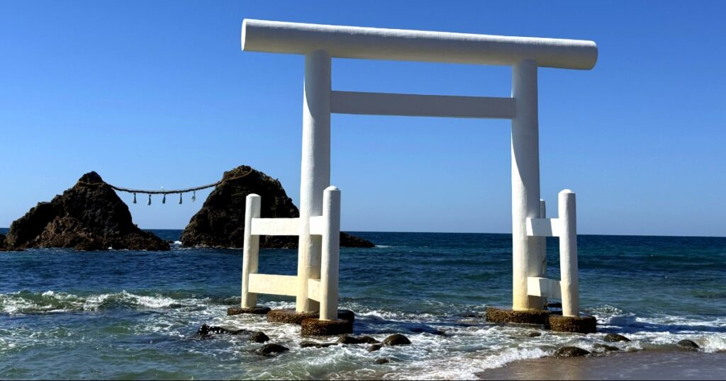 White torii gate stands in ocean waves at Japanese coastal shrine beach on Itoshima Island.