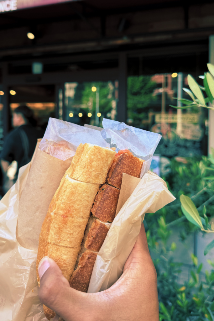 Hand holding mentaiko bread sandwich in front of Fukuoka bakery storefront.