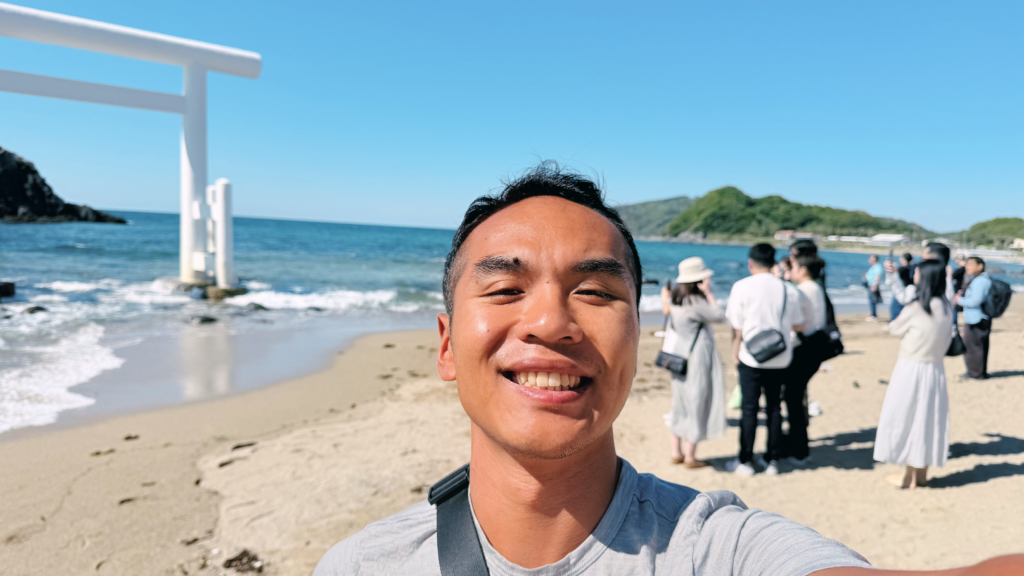 Man taking selfie at Itoshima Island beach with white torii gate and tourists in the background.