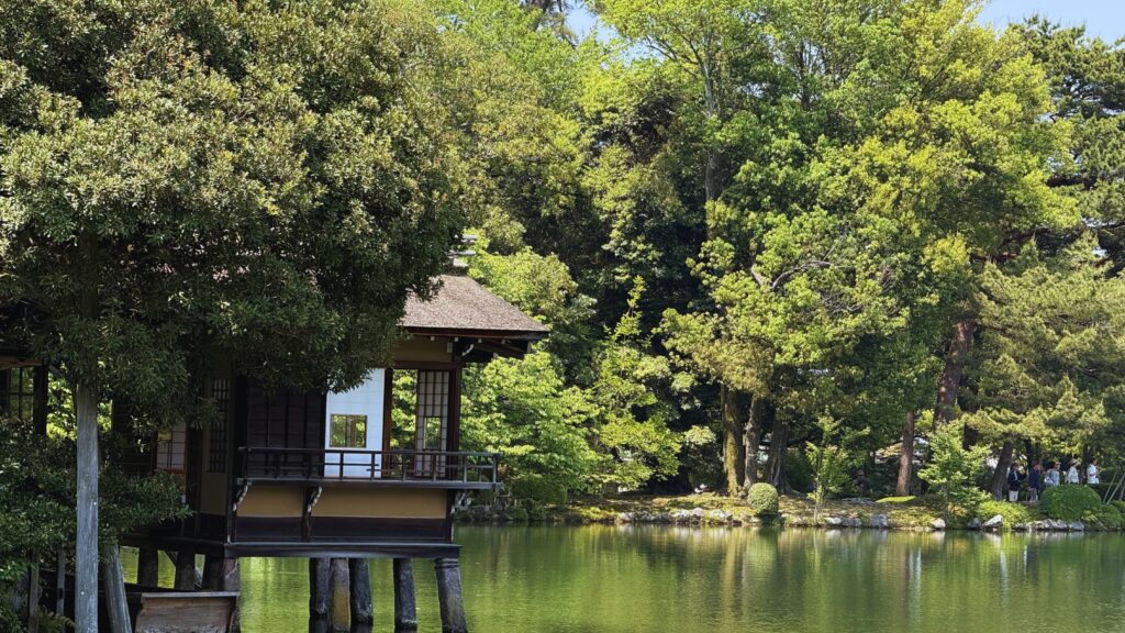 Traditional teahouse on stilts over a serene pond surrounded by lush green trees in Kenroku-en garden, Kanazawa