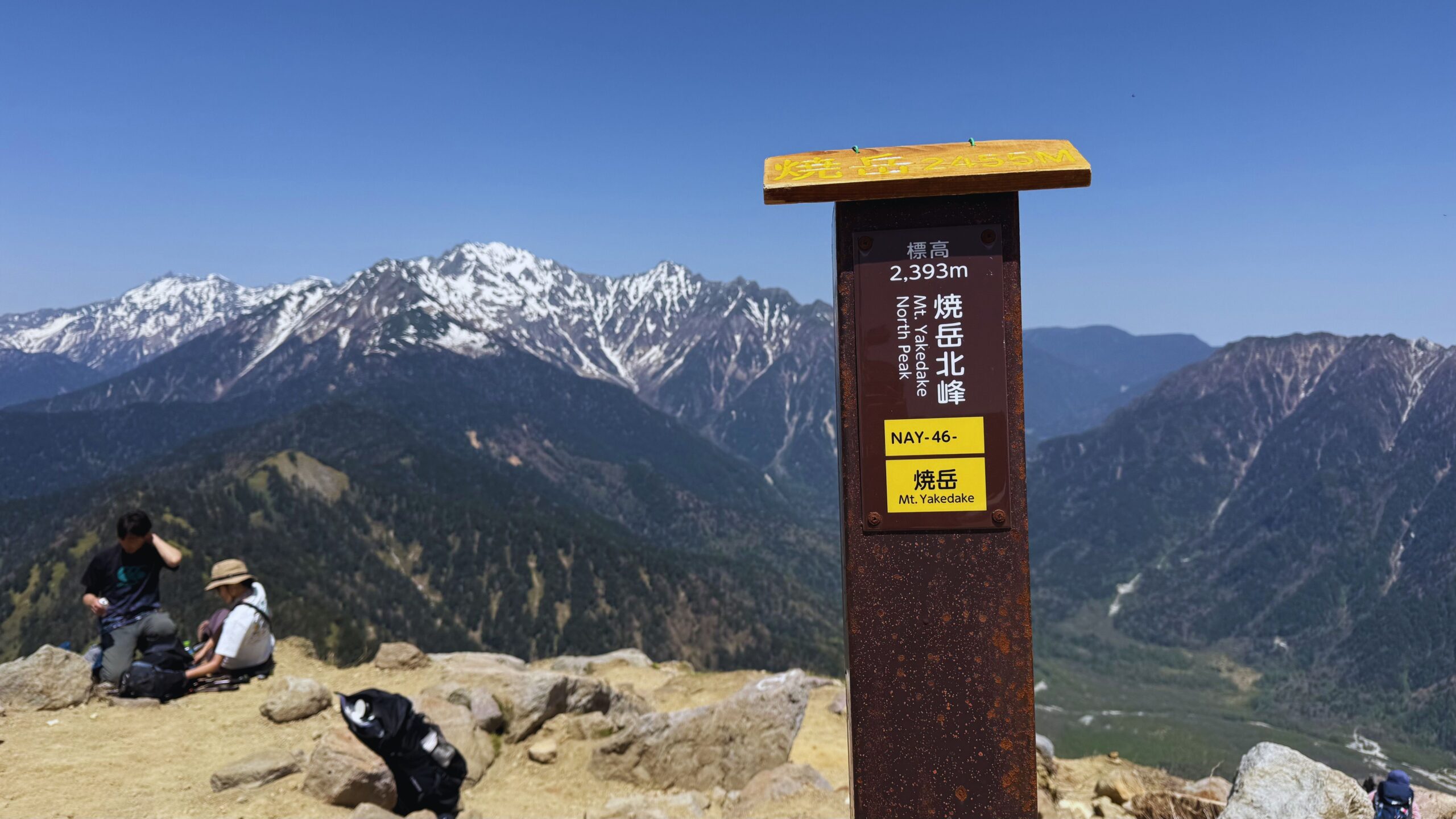 Summit sign at 2,393 meters on Mount Yakedake with Northern Alps panorama stretching behind it