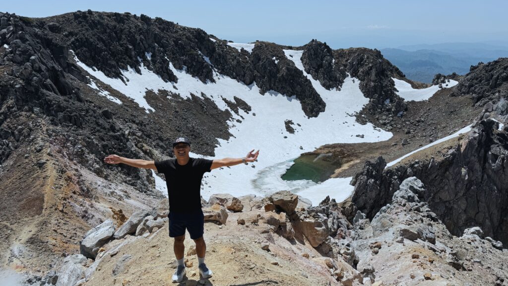 Traveler standing on the rocky summit of Mount Yakedake with snow fields and a crater lake visible below