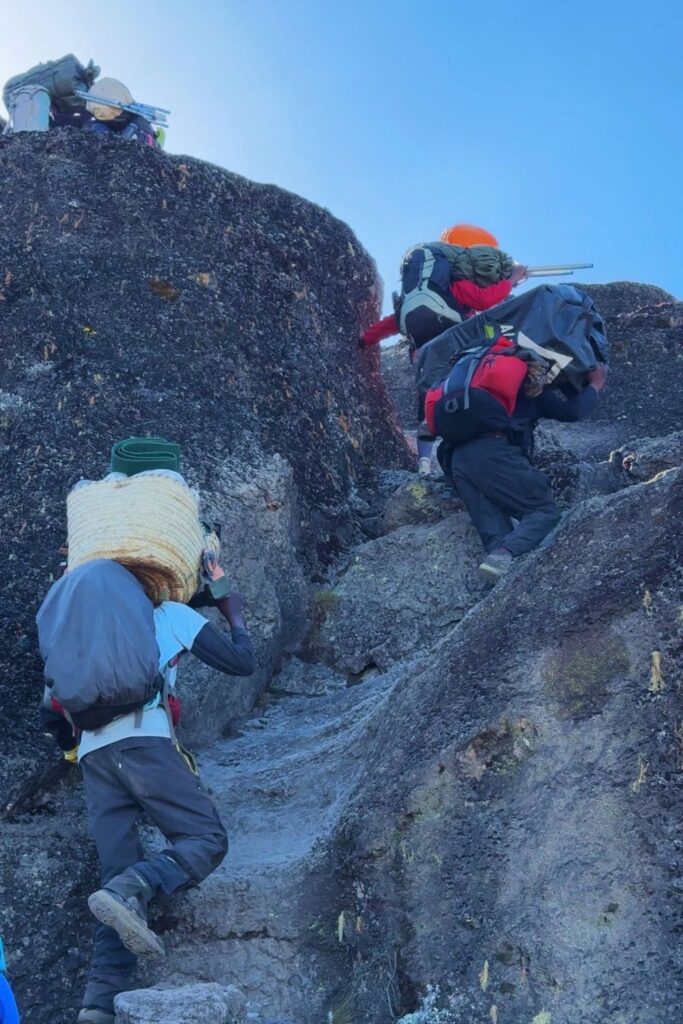 Porters scrambling up the rocky Barranco Wall while carrying heavy loads on their backs