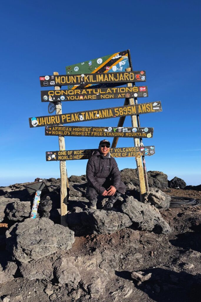 Crouched in front of the Uhuru Peak summit sign at 5,895 meters, the top of Kilimanjaro