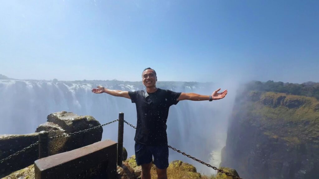 A man smiling with his arms spread wide at a viewpoint on the edge of the gorge, with Victoria Falls and its spray behind him