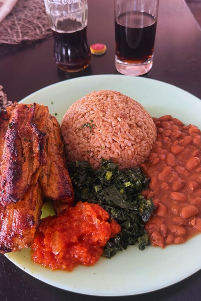 A plate of grilled meat, rice, beans, cooked greens, and tomato relish, with two glasses of cola at a restaurant in Victoria Falls