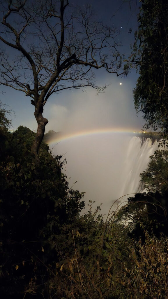 A faint lunar rainbow arching over the mist of Victoria Falls at night, with a bare tree and stars in the sky
