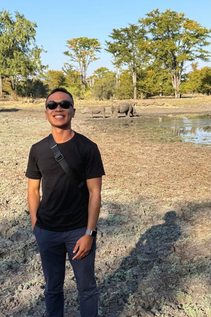 A man smiling in sunglasses on dry grassland, with a white rhino grazing near a waterhole behind him
