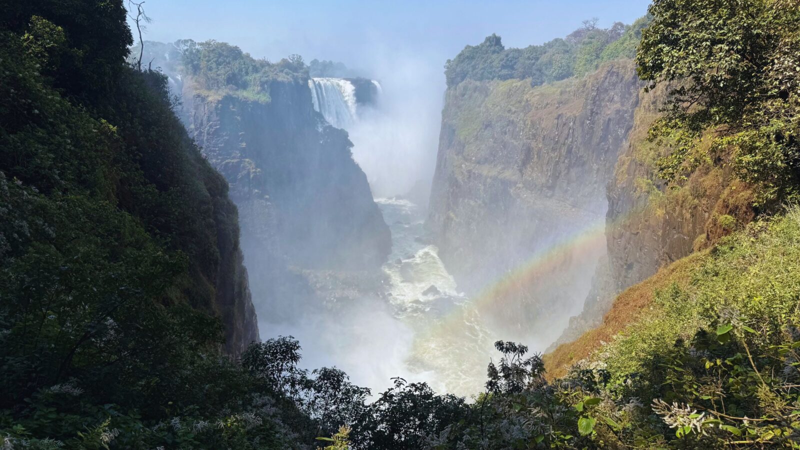 Rainbow arching over the spray in the gorge below Victoria Falls, framed by green trees on the cliffs