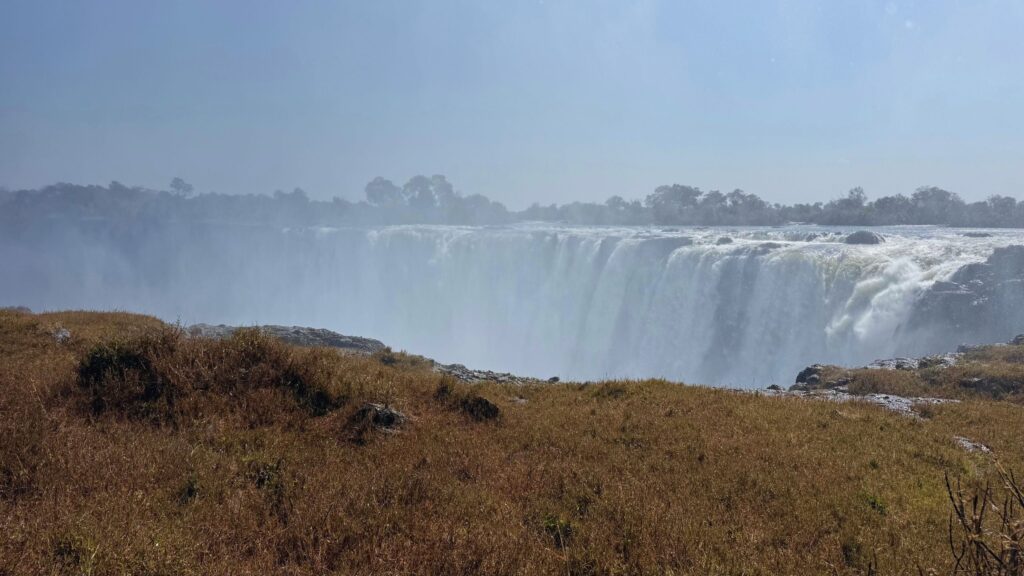 Victoria Falls pouring over the cliff in a cloud of mist, seen across dry grass at the edge of the gorge