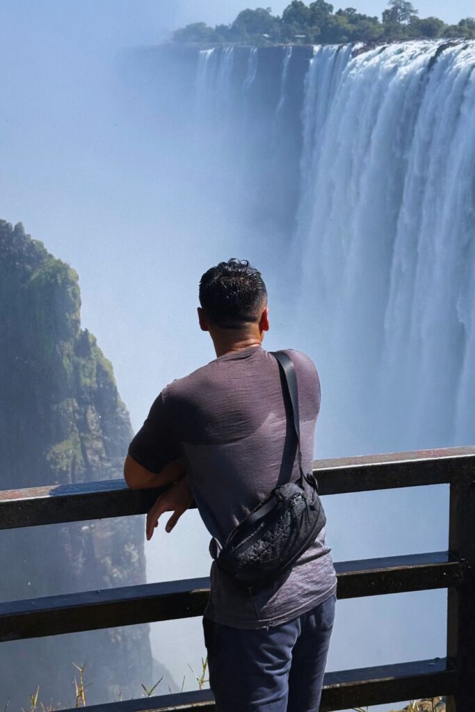 A man leaning on a railing with his back to the camera, looking at Victoria Falls pouring into the gorge