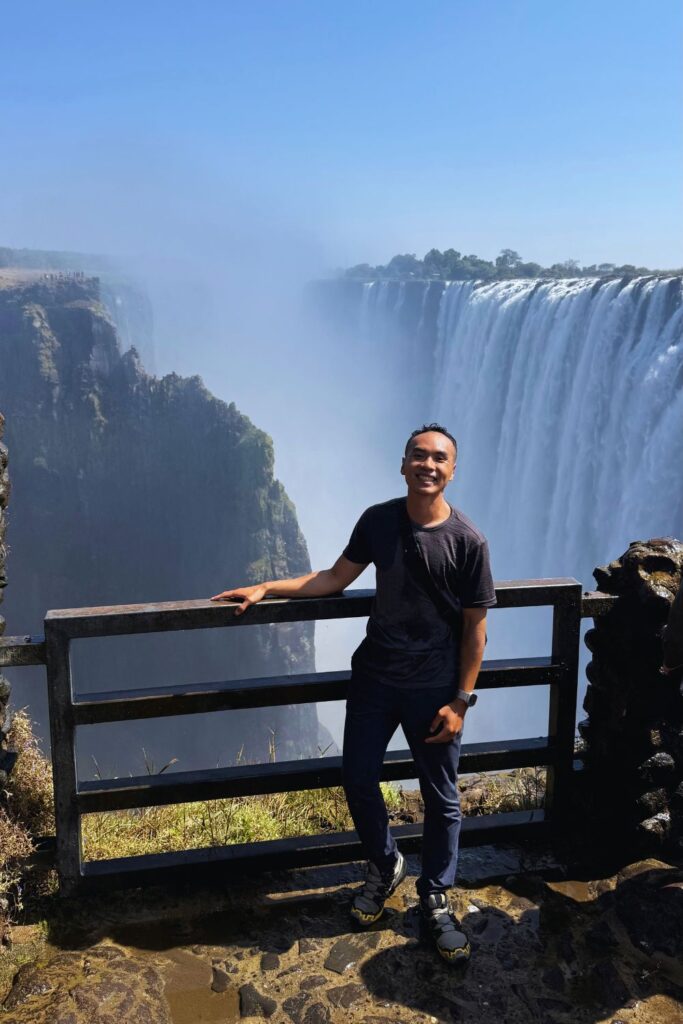 A man smiling at a viewpoint railing with the curtain of Victoria Falls and a clear blue sky behind him