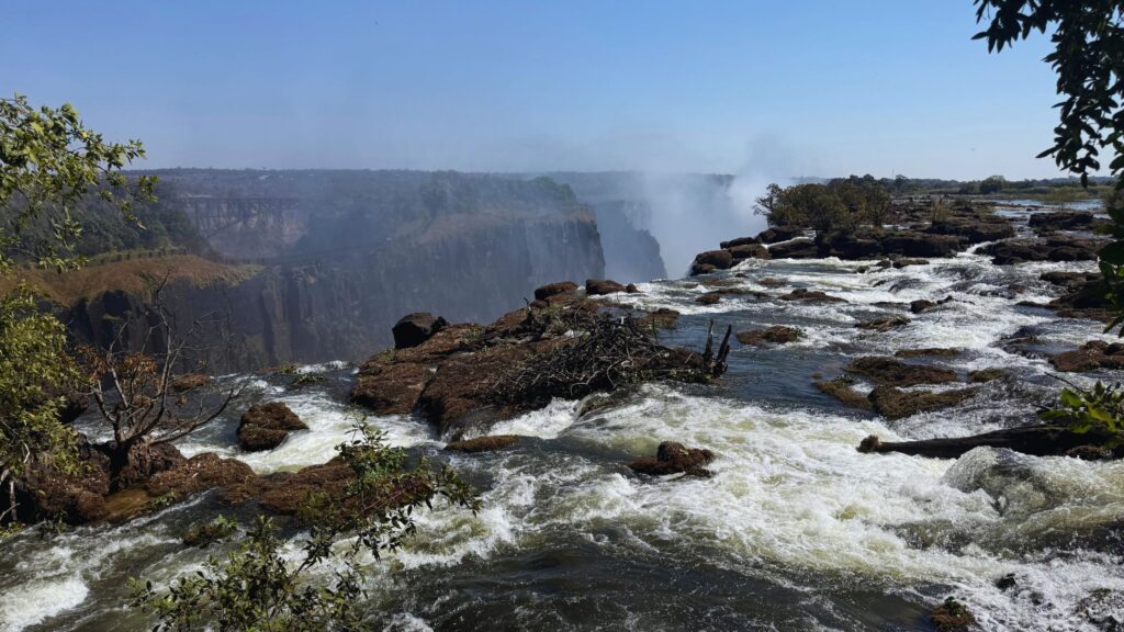 Zambezi River rapids rushing over rocks toward the edge of Victoria Falls, with spray rising from the gorge and Victoria Falls Bridge in the distance