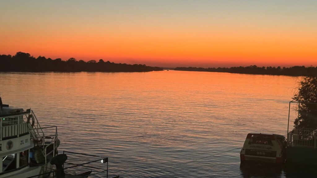 Orange sunset over the Zambezi River with boats docked along the shore near Victoria Falls