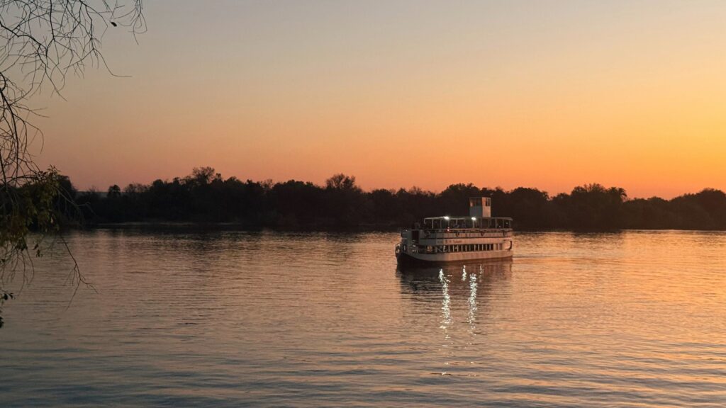 A sunset cruise boat on the Zambezi River at dusk, with trees silhouetted against an orange sky