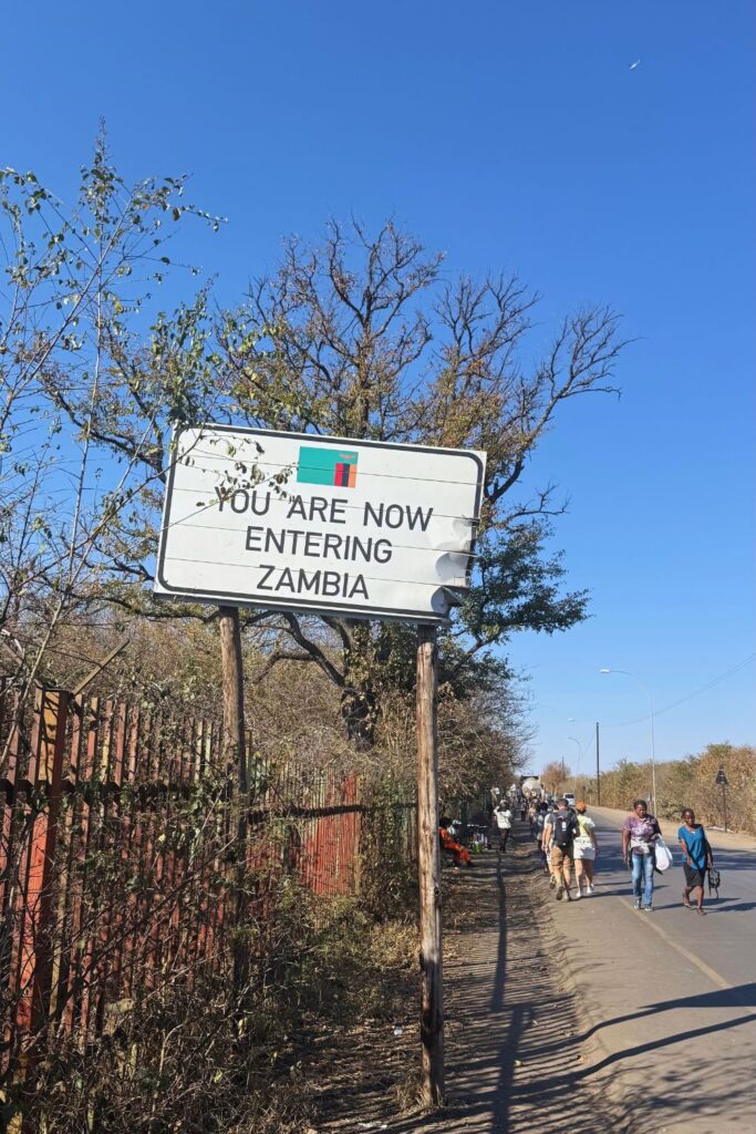 You Are Now Entering Zambia sign with the Zambian flag as people walk toward the Victoria Falls border
